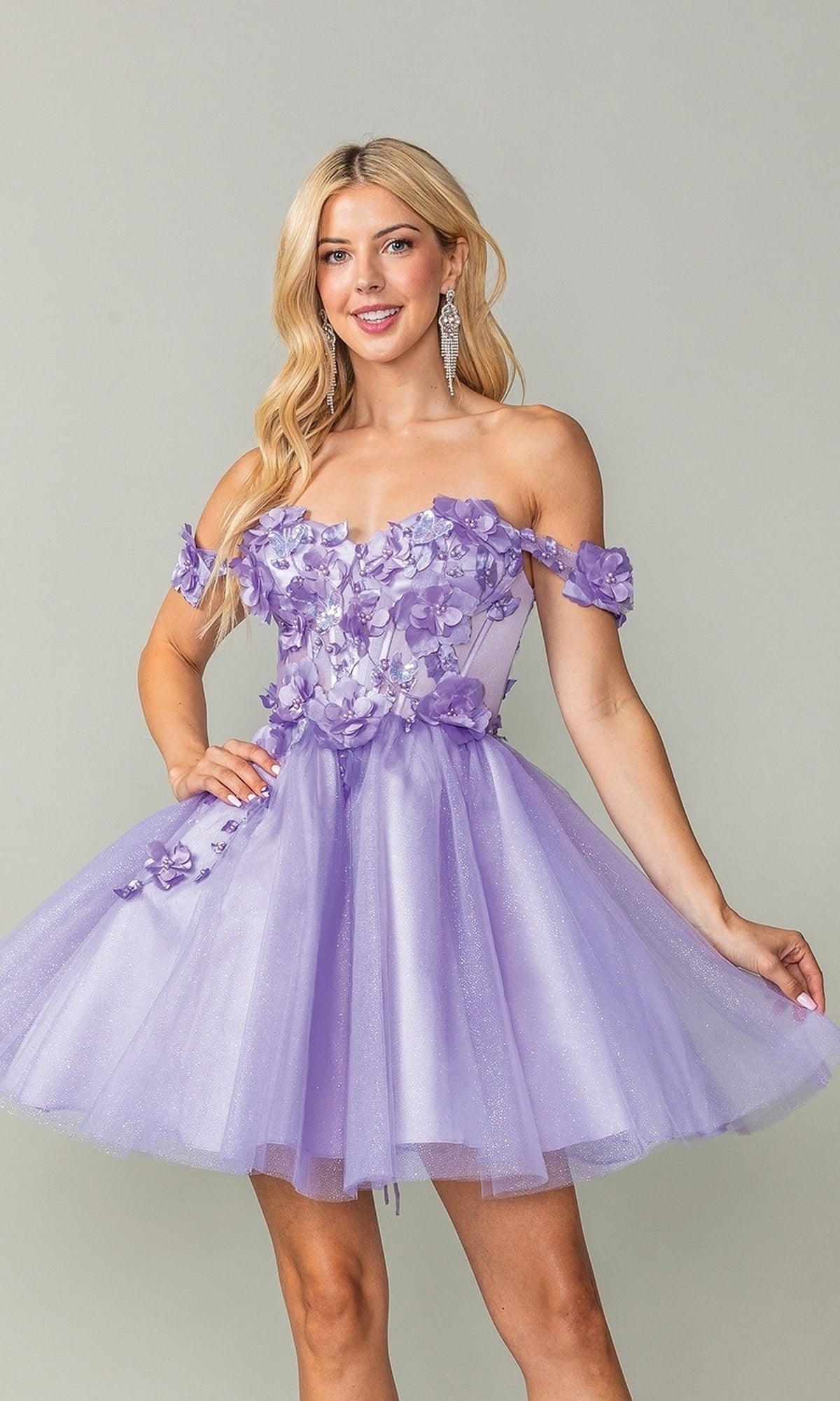 A woman models the Off-Shoulder Short Babydoll Homecoming Dress 3388, featuring floral appliqués and a tulle skirt, in front of a plain gray background.